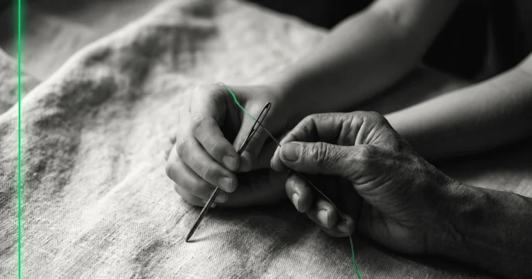 Deux mains enfilent un fil vert dans le chas d'une grosse aiguille ancienne, sur un lin écru, en noir et blanc.