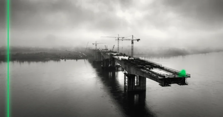Un pont inachevé s'arrête au-dessus d'un large fleuve dans la brume, grues à l'arrêt.