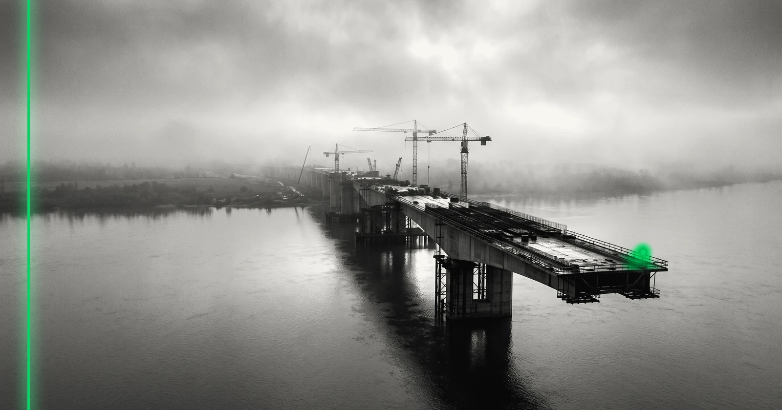 Un pont inachevé s'arrête au-dessus d'un large fleuve dans la brume, grues à l'arrêt.