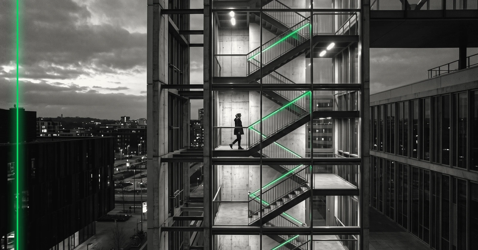 Une silhouette gravit une cage d'escalier entièrement vitrée, dont toute la structure est visible au crépuscule.